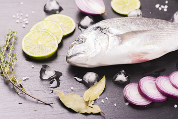 Fresh raw dorada fish with lime, red onion, spices and herbs over black stone background. Selective focus. Top view