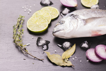 Fresh raw dorada fish with lime, red onion, spices and herbs over black stone background. Selective focus. Top view