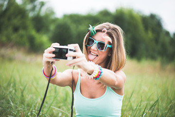 Young woman taking self portrait outdoor, wearing funny sunglasses and holding a compact camera