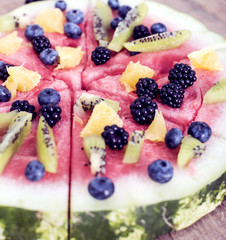 colorful tropical fruit watermelon pizza topped with kiwifruit, blueberries, orange and fresh berries cut into segments on a rustic wooden board