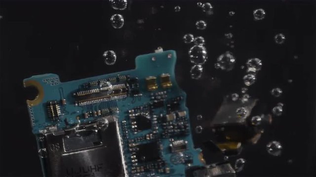 A torn apart cell phone slowly floats down in a clear substance with bubbles
