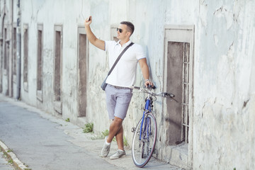 Young man standing on the old town street with his bicycle beside him and taking a photo with his smart phone