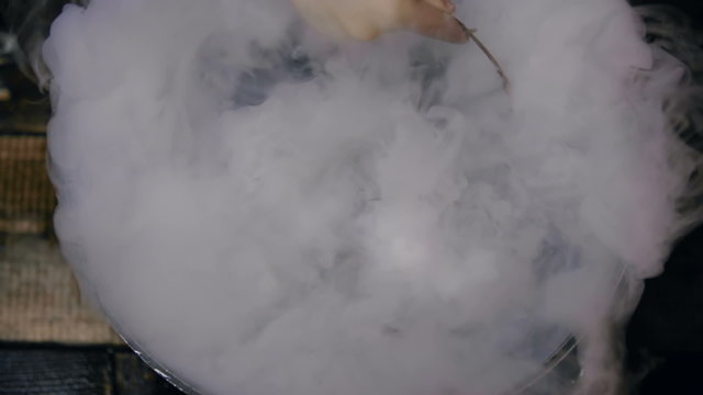 Hands of little witch putting ingredients into smoky cauldron with boiling potion 