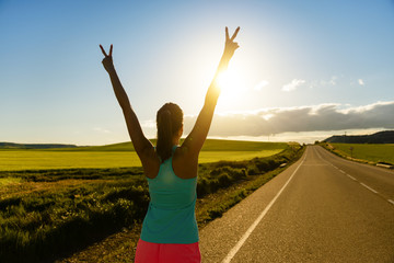 Woman celebrating running and training success on countryside road during sunset or sunrise. Female runner raising arms towards the sun.