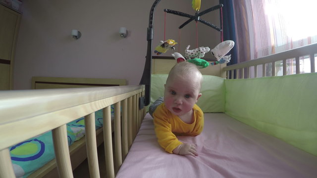 Infant child have fun in bed near sun lighted window. Playful interested baby turn over under of spinning carousel over wooden bed crib. Wide angle shot. 4K UHD.
