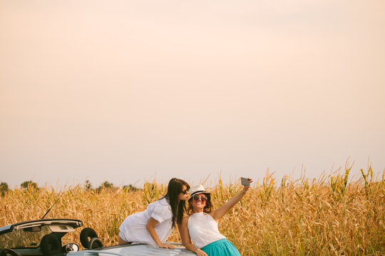 Two female friends beside cabriolet taking selfies with smartphone