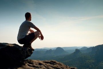 Naklejka na ściany i meble Young man in black sports pants and grey shirt is sitting on cliff's edge and looking to misty valley bellow