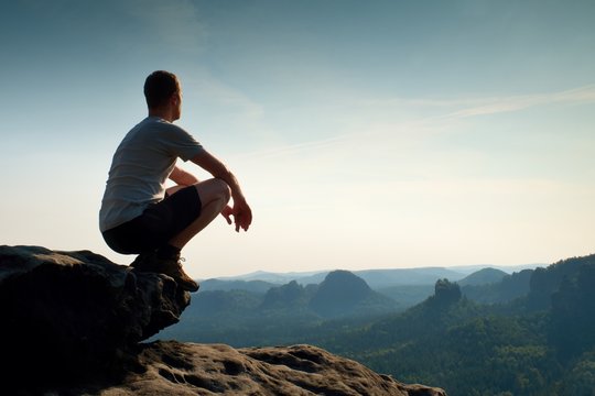 Young Man In Black Sports Pants And Grey Shirt  Is Sitting On Cliff's Edge And Looking To Misty Valley Bellow