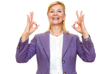 Happy smiling old senior woman 60-65 years with white teeth, shows gesture ok on two hands. In suit with big blue eyes, isolated on white background, Positive human emotion, facial expression