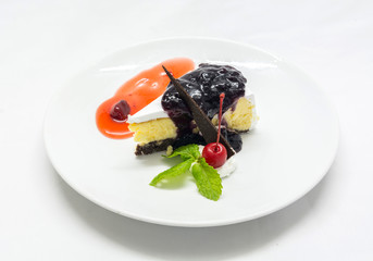 Cheese cake dressing with white cream, blue berry jam, red berry jam, red cherry, mint leaves and chocolate on a plate on white background