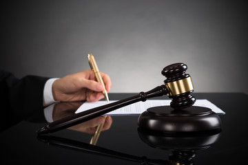 Close-up Of Judge Writing On Paper And Gavel At Desk