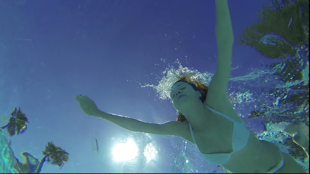 Slim pretty girl in a white bathing suit swims underwater in the sunshine.
