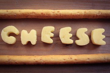 The word cheese written in chunks of cheddar on a wooden surface with sesame seed bread sticks