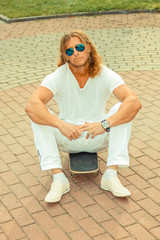 muscular long-haired guy in sunglasses with skateboard. Urban scene. Outdoor lifestyle portrait