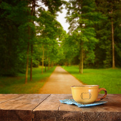 front image of coffee cup over wooden table and autumn leaves in front of autumnal forest 
