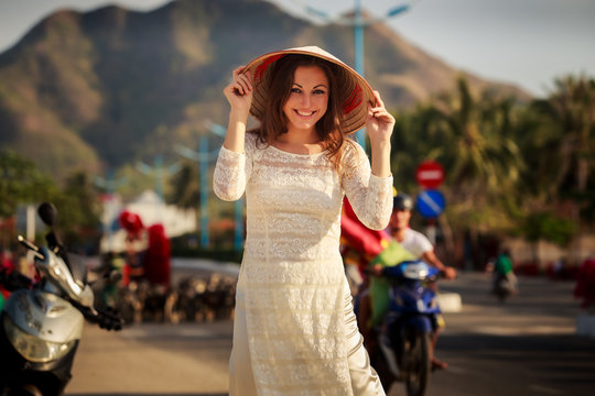 girl in Vietnamese dress touches hat against defocused scooters
