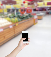 Hand holding mobile phone with Vegetables and fruit on shelf in