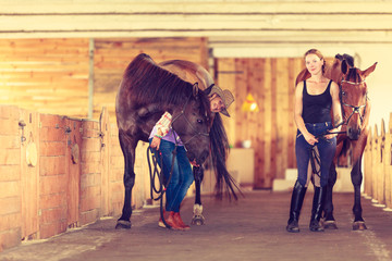 Cowgirl and young woman in stable with horses.