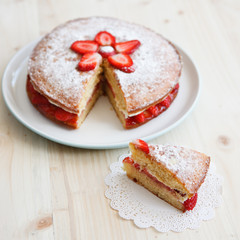 Victoria sponge cake with strawberries, jam and whipped cream with a cut out piece on a wooden table