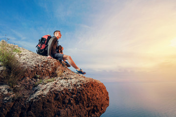 A young man hiker  with a backpack hiking looking through binoculars sitting on a rock mountain over the sea.