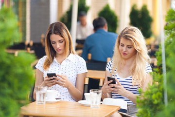 Two young women using mobile phones while drinking coffee in a cafe