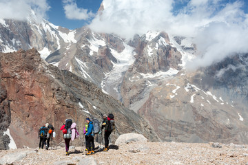Backpackers Team Walking on Rocky Trail Group of People in Sporty Style Clothing with Backpacks and Walking Poles Mountain Landscape with Blue Sky and Clouds on Background