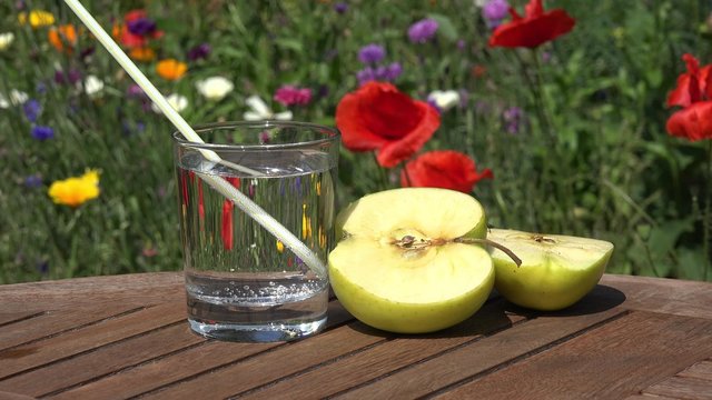 Glass Water and Apple on table in the garden in front of summer flowers