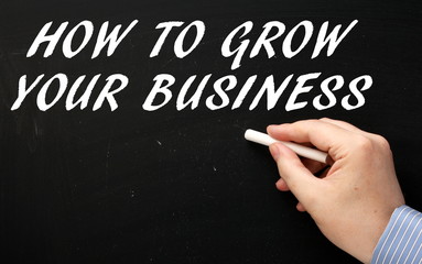 Hand writing the phrase How To Grow Your Business in white text on a blackboard using a stick of chalk