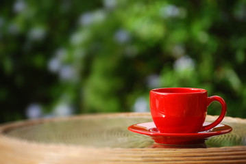 Photograph of a vibrant red cappuccino mug, on a wooden table's glass, with glorious green bokeh in the background.