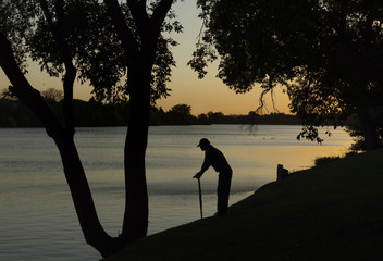 horizontal silhouette of an old man leaning on walking cane looking out across the lake at sunset with trees lining the banks on a summer evening.