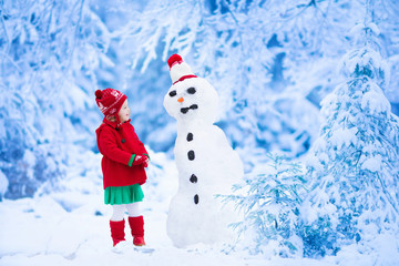 Funny little toddler girl in a red knitted Nordic hat and warm coat playing with a snow man. Kids...