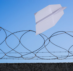Papierflieger mit Textfreiraum im Steigflug über Absperrung aus Beton und Stacheldraht
