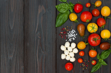 Cherry tomatoes of various color, mozzarella, basil leaves, spices from above over dark wooden table. Italian caprese salad recipe ingredients. Top view, free text copy space