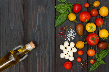 Cherry tomatoes of various color, mozzarella, basil leaves, spices and olive oil from above over dark wooden table. Italian caprese salad recipe ingredients. Top view, free text copy space