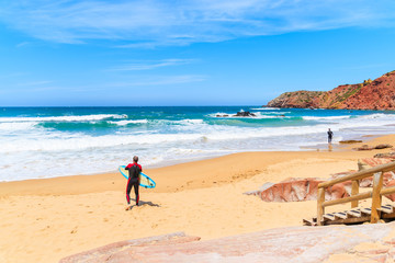 Surfer standing on Praia do Amado beach with ocean waves hitting shore, Algarve region, Portugal