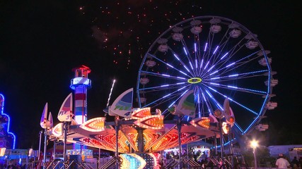 Fireworks explode in the night sky behind a ferris wheel at a carnival or state fair. - Powered by Adobe