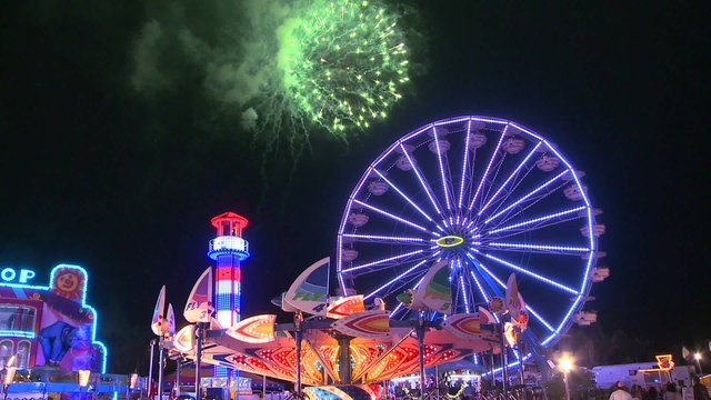 Fireworks explode in the night sky behind a ferris wheel at a carnival or state fair.