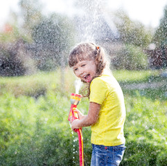 Cheerful kid watering plants from hose spray in garden at