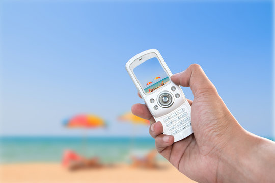 Human hand holding vintage phone shooting picture at the sea with image of blurred colorful umbrella