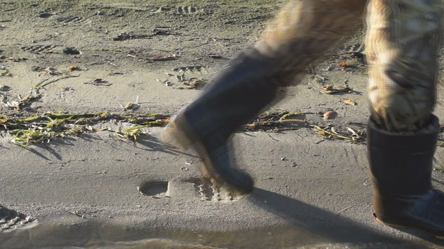 A Man With Brown,  Camouflage, Rubber Waders Walks Along The Beach Of A Lake Shore Leaving Footprints On The Shore Of The Sand Beach.