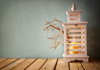 low key image of white wooden vintage lantern with burning candle and tree branches on wooden table. retro filtered image

