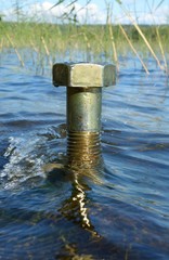Single big metal nut stands in the water with green reeds on the background in a one of the  Finland's clean water lakes with ecology, nature protection and green technology concept idea.