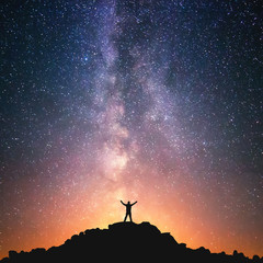 Man and the Universe. A person is standing on the top of the hill next to the Milky Way galaxy with his hands raised to the air. - obrazy, fototapety, plakaty