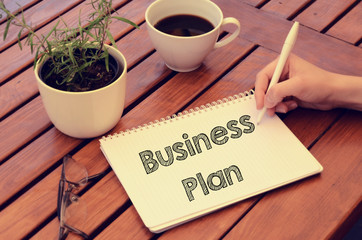 Woman hand writing in notebook Business Plan on wooden table wit