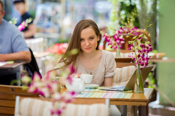 Young beautiful woman sitting in cafe and drinking coffee. Working with the laptop on terrace.
