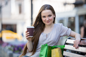 Young beautiful woman sitting on a bench on the street with coffee cup in hand and shopping bags. Drinking coffee and smiling.
