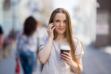 Young woman walking on the street and drinking coffee. She is talking on the phone with a take away cup of coffee
