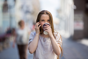 Young woman walking on the street and drinking coffee. She is talking on the phone with a take away cup of coffee
