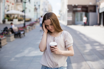 Young woman standing on the street suffering from pain. She has a take away cup of coffee and touch her temple
