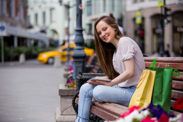 Young beautiful woman sitting on a bench with digital tablet next to the shopping bags. Relaxing and using the device with happy smile
